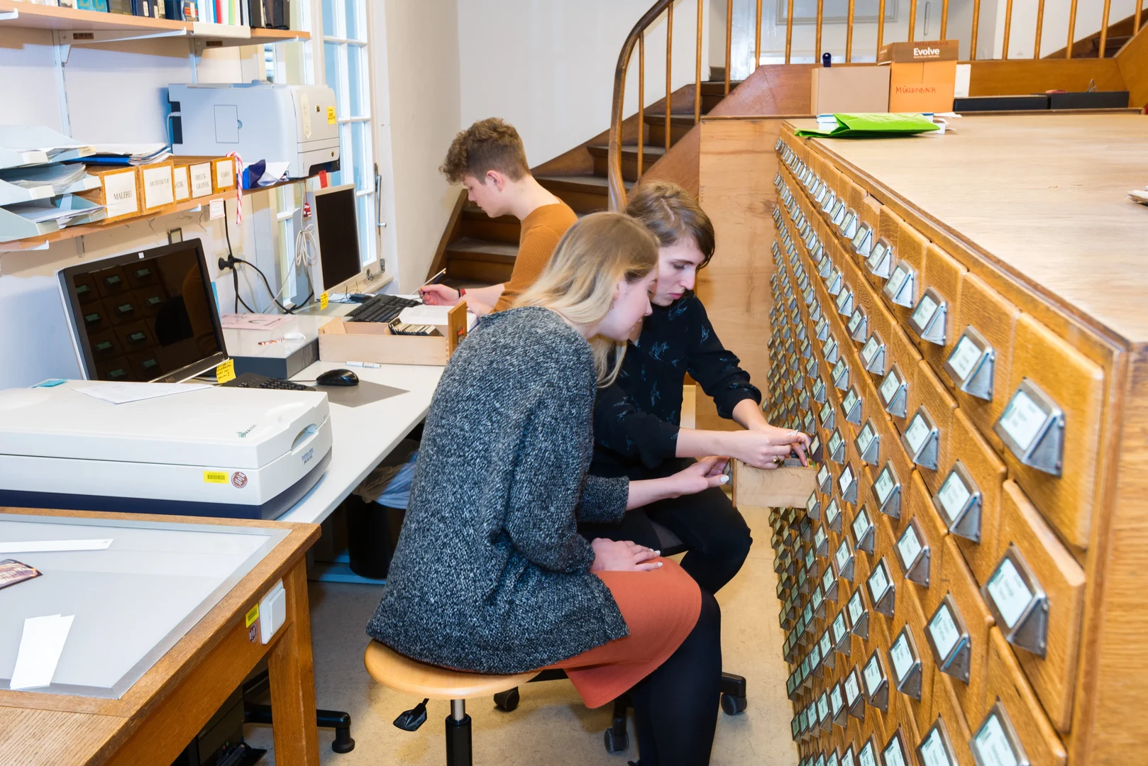 Eine Wissenschaftlerin und ein Wissenschaftler arbeiten hinter einer Glasfassade und mischen Chemikalien mit Großgeräten.