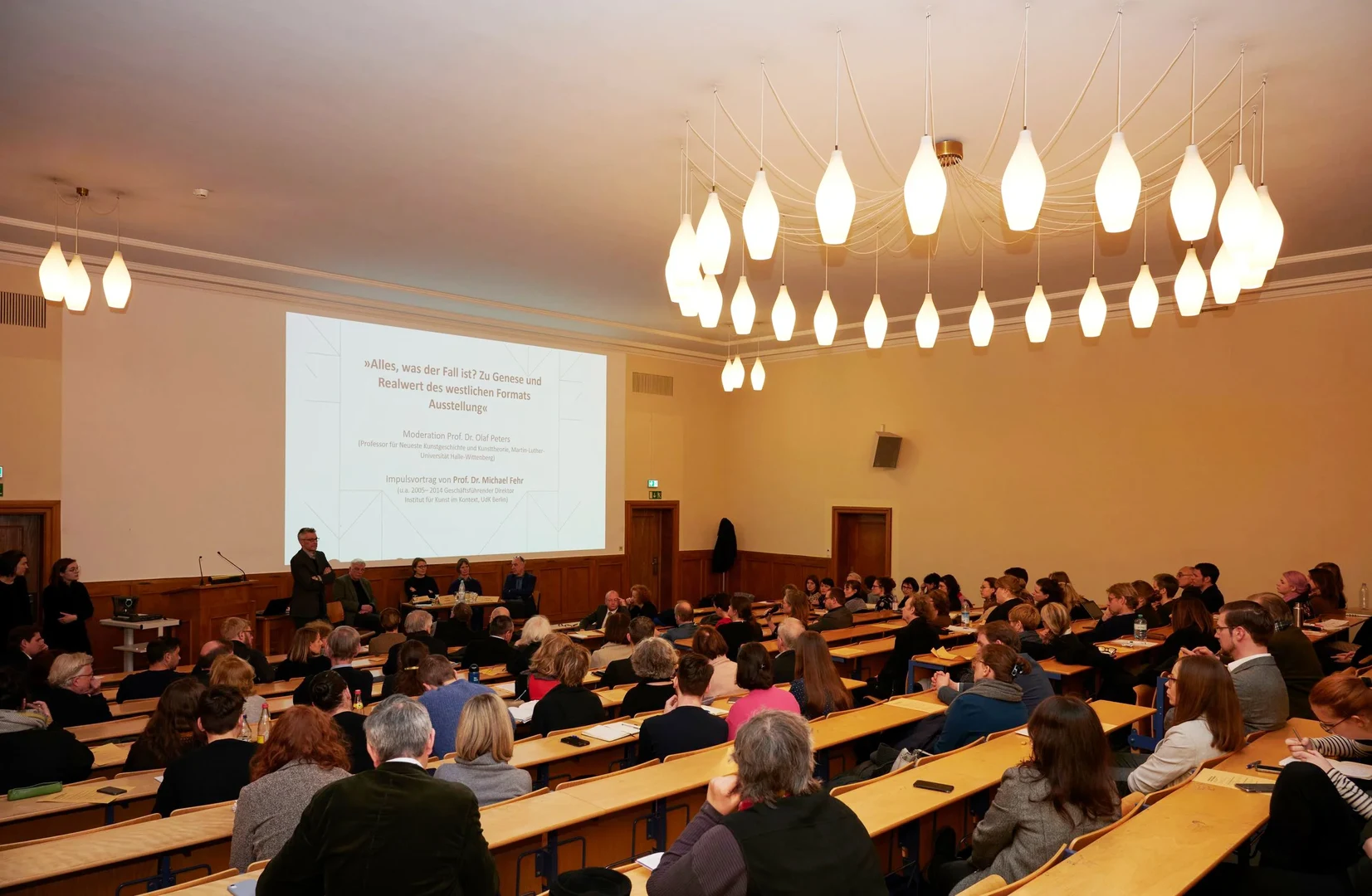 Eine Wissenschaftlerin und ein Wissenschaftler arbeiten hinter einer Glasfassade und mischen Chemikalien mit Großgeräten.