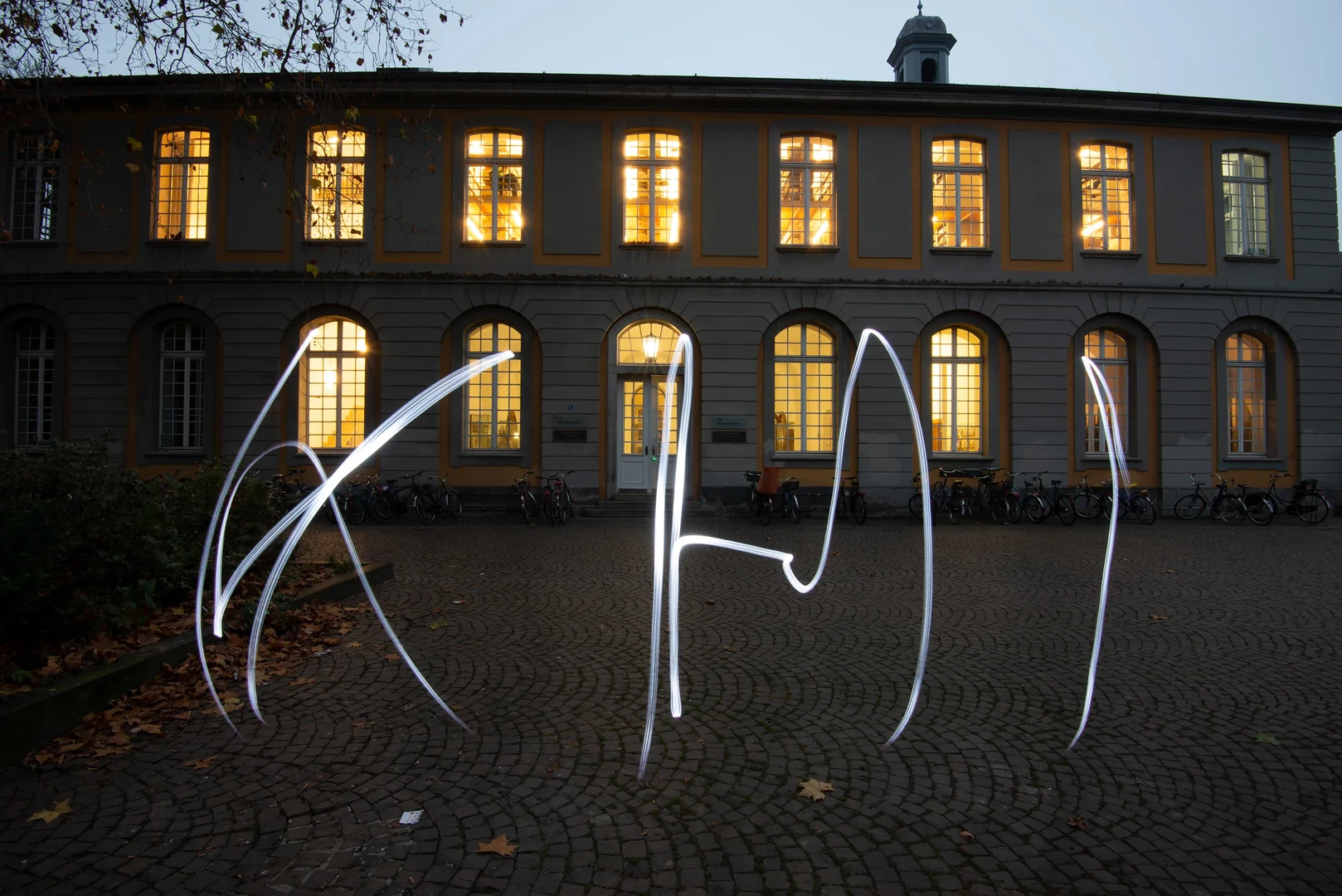 Eine Wissenschaftlerin und ein Wissenschaftler arbeiten hinter einer Glasfassade und mischen Chemikalien mit Großgeräten.
